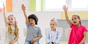 four yound students sitting in a classroom; three have their hand raised.